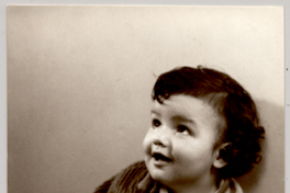 [Niñito moreno, regordete, con pantalons oscuros y un chaleco con guardas claras con cierre, sentado sobre una colchoneta forrada con brocato y con dos juguetes]