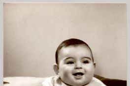 [Bebé moreno con un traje de lana color blanco sentado sobre una colchoneta de brocato con dos plumones en sus espaldas y un juguete en sus manos]