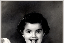 [Niña posando con un vestido blanco de mangas cortas y una rosa en la espalda y tiene un juguete en sus manos]