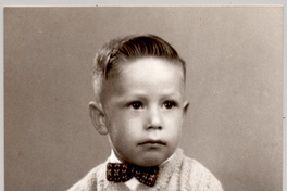 [Niño rubio sentado sobre cubrecama de brocato con pantalones cortos, chaleco y un corbatín, con las manos apoyadas en sus muslos]