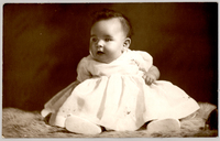 [Bebé con amplio vestido blanco, cara regordeta, sentada sobre un felpudo]
