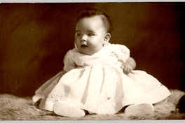 [Bebé con amplio vestido blanco, cara regordeta, sentada sobre un felpudo]