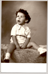 [Niñito de pantalón corto, camisa, sandalias y soquetes blancos, pelo ondulado peinado hacia atrás, sentado sobre un felpudo]