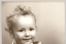 [Niñito rubio de frente amplia, vestido con un traje pantalón corto color oscuro y blusa manga corta blanca con un juguete en sus manos sentado sobre un mueble forrado con brocato]