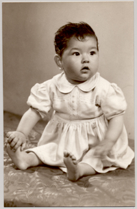 [Niñita con rasgos asiáticos con un vestido claro recogido con cuello, abotonado y mangas englobasdas sentada sobre una colchoneta de brocato y a pie pelado]