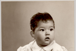 [Niñita con rasgos asiáticos con un vestido claro recogido con cuello, abotonado y mangas englobasdas sentada sobre una colchoneta de brocato y a pie pelado]