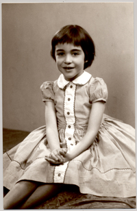 [Niña sentada con un amplio vestido de tela recogido con una guarda blanca con bordados de colores y con ls manos puestas una sobre la otra]