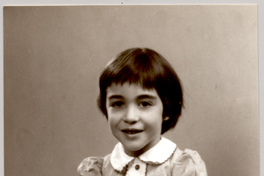 [Niña sentada con un amplio vestido de tela recogido con una guarda blanca con bordados de colores y con ls manos puestas una sobre la otra]