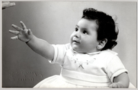 [Niñita de vestido vaporoso, chalas y calcetines blancos sentada sobre una colchoneta de brocato y en sus manos tiene al Pato Donald]