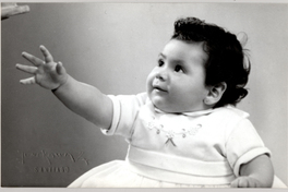 [Niñita de vestido vaporoso, chalas y calcetines blancos sentada sobre una colchoneta de brocato y en sus manos tiene al Pato Donald]