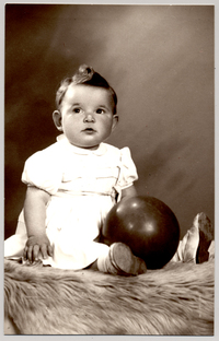 [Guagua rubia con el pelo tomado hacia arriba, vestido mangas cortas, con una pelota y sentada sobre un felpudo]