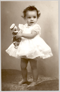 [Niñita con un vestido de holganza con fondo blanco, con un juguete en sus manos y de pie sobre una colchoneta forrada en brocato]