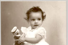 [Niñita con un vestido de holganza con fondo blanco, con un juguete en sus manos y de pie sobre una colchoneta forrada en brocato]