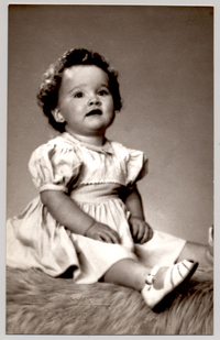 [Niñita de pelo crespo sentada sobre un felpudo con un vestido blanco recogido, una cinta de adorno en la pechera, mangas englobadas y chalas blancas]