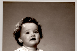 [Niñita de pelo crespo sentada sobre un felpudo con un vestido blanco recogido, una cinta de adorno en la pechera, mangas englobadas y chalas blancas]