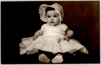 [Bebé sentada con un vestido muy amplio color blanco y un sombrero del mismo color y chalas]