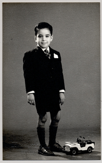 [Niño vestido con terno oscuro de pantalón corto, corbata, pañuelo blanco en el bolsillo y un camión de juguete en el suelo]