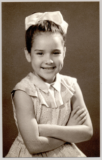 [Niña de unos ocho años, posando sonriente con un vestido de broderíblanco, con vuelos en el cuello, una cinta en el pelo y de brazos cruzados]