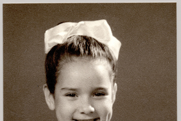 [Niña de unos ocho años, posando sonriente con un vestido de broderíblanco, con vuelos en el cuello, una cinta en el pelo y de brazos cruzados]