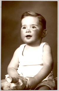 [Bebé rubio cara rosasea polera blanca sin mangas con un juguete en sus manos y sonriendo]