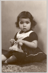 [Niñita morena de pelo corto con chasquilla sentada sobre una colchoneta forrada en brocato con un vestido oscuro y una cinta de broderí en el cuello y con un teléfono de juguete en sus manos]