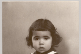 [Niñita morena de pelo corto con chasquilla sentada sobre una colchoneta forrada en brocato con un vestido oscuro y una cinta de broderí en el cuello y con un teléfono de juguete en sus manos]