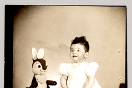 [Niña de vestido blanco con mangas englobadas sentada con un felpudo y con un conejo de peluche]