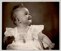 [Guagua sonriente, rubia de pelo rizado, con un vestido de verano de gasa blanco, una medalla colgando en el cuello y una pulsera en su mano izquierda]