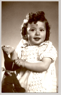 [Niñita de pelo rizado, vestido blanco con un juguete en sus manos]