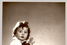 [Niñita de pelo rizado, vestido blanco abrazada a un juguete sentada sobre un felpudo]