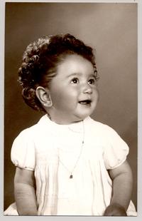 [Niñita de pelo rizado peinado hacia atrás vestido blanco mangas cortas y una cadena colgando en el cuello]