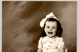 [Niñita sentada sobre un felpudo con un vestido repolludo con flores, sandalias blancas con soquetes, una cinta en el pelo y con un juguete en sus manos]