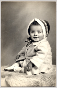 [Niñita con abrigo y un gorro de lana de colores sentada sobre un felpudo]