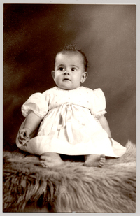 [Niñita de vestido recogido con una cinta blanca, mangas cortas englobadas y descalza sentada sobre un felpudo]