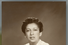 [Mujer morena de pelo corto, blusa blanca manga corta y un prendedor en el cuello, sentada en un sillón de estudio]