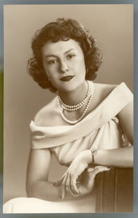 [Mujer posando de medio cuerpo con un vestido escotado y un collar blanco de tres vueltas, apoyada en un sillón de estudio]