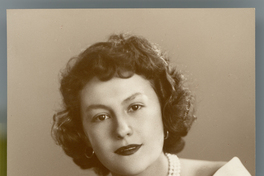 [Mujer posando de medio cuerpo con un vestido escotado y un collar blanco de tres vueltas, apoyada en un sillón de estudio]