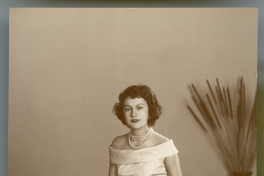 [Mujer posando con un vestido amplio y largo, hombros al desnudo, collar blanco de tres vueltas, sentada en una tarima y al fondo un macetero con espigas]