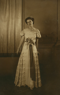 [Mujer joven de vestido largo con una gran rosa en la cintura y un collar de flores en el cuello]