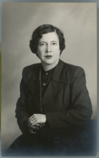 [Mujer de traje dos piezas y blusa abotonada negra, uñas pintadas rojas, sentada con las manos una encima de la otra]