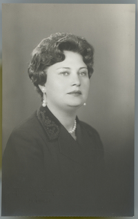 [Mujer adulta de pelo corto, regordeta, tez y ojos claro, abrigo oscuro bordado en la solapa, aros y collar de perlas blancas]