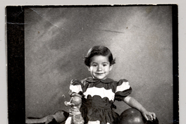 [Niñita de vestido rojo y blanco, con una pelota y un pato de juguete en sus manos mirando de frente]