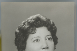 [Mujer adulta de pelo corto ondulado, labios rojos, sweater claro y aros y colar de perlas blancas]