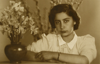 [Mujer joven, vestida de verano y apoyada en un mueble. En frente tiene un macetero con flores blancas y a sus espaldas se ve unos brotes de ramas largas]