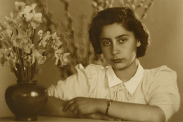 [Mujer joven, vestida de verano y apoyada en un mueble. En frente tiene un macetero con flores blancas y a sus espaldas se ve unos brotes de ramas largas]