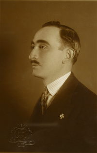 [Hombre posando de perfil, con traje oscuro y un prendedor en la solapa del vestón, de estilo elegante]