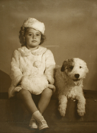 [Niña sentada sobre una colchoneta vestida con un abrigo y gorro de piel color blanca acompañada de un perro blanco con una mancha en su ojo derecho]