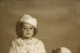 [Niña sentada sobre una colchoneta vestida con un abrigo y gorro de piel color blanca acompañada de un perro blanco con una mancha en su ojo derecho]