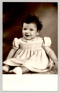 [Niñita de ojos claros, pelo rizado, vestido con mangas englobadas, pulsera en su mano izquierda posando sobre un felpudo]
