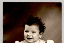 [Niñita de ojos claros, pelo rizado, vestido con mangas englobadas, pulsera en su mano izquierda posando sobre un felpudo]